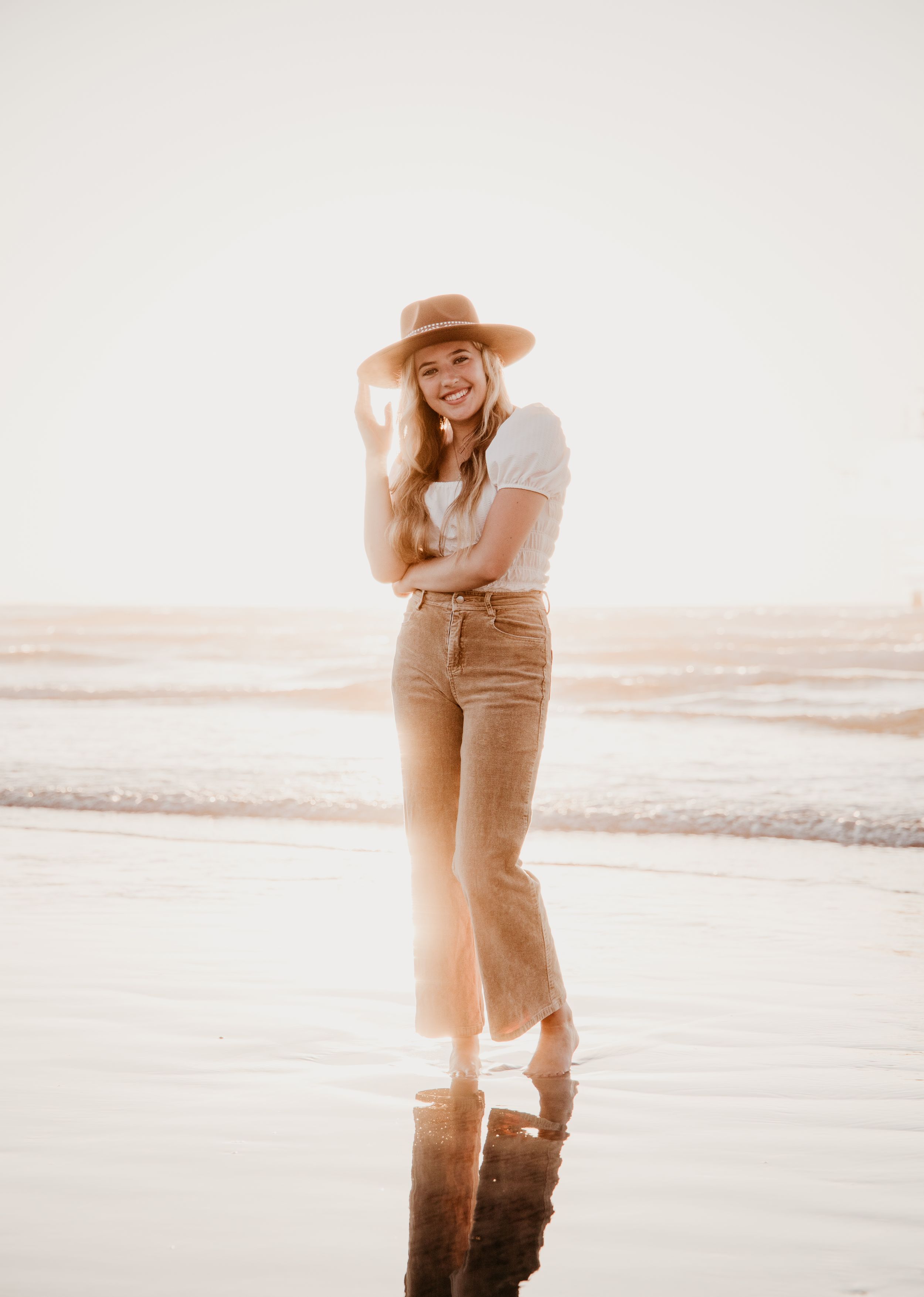 Person in casual attire and sun hat poses on reflective beach at golden hour.