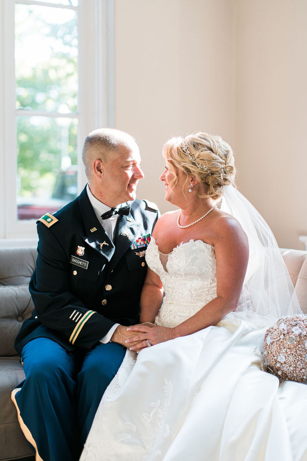 Military groom and bride in white dress share tender moment on elegant sofa.