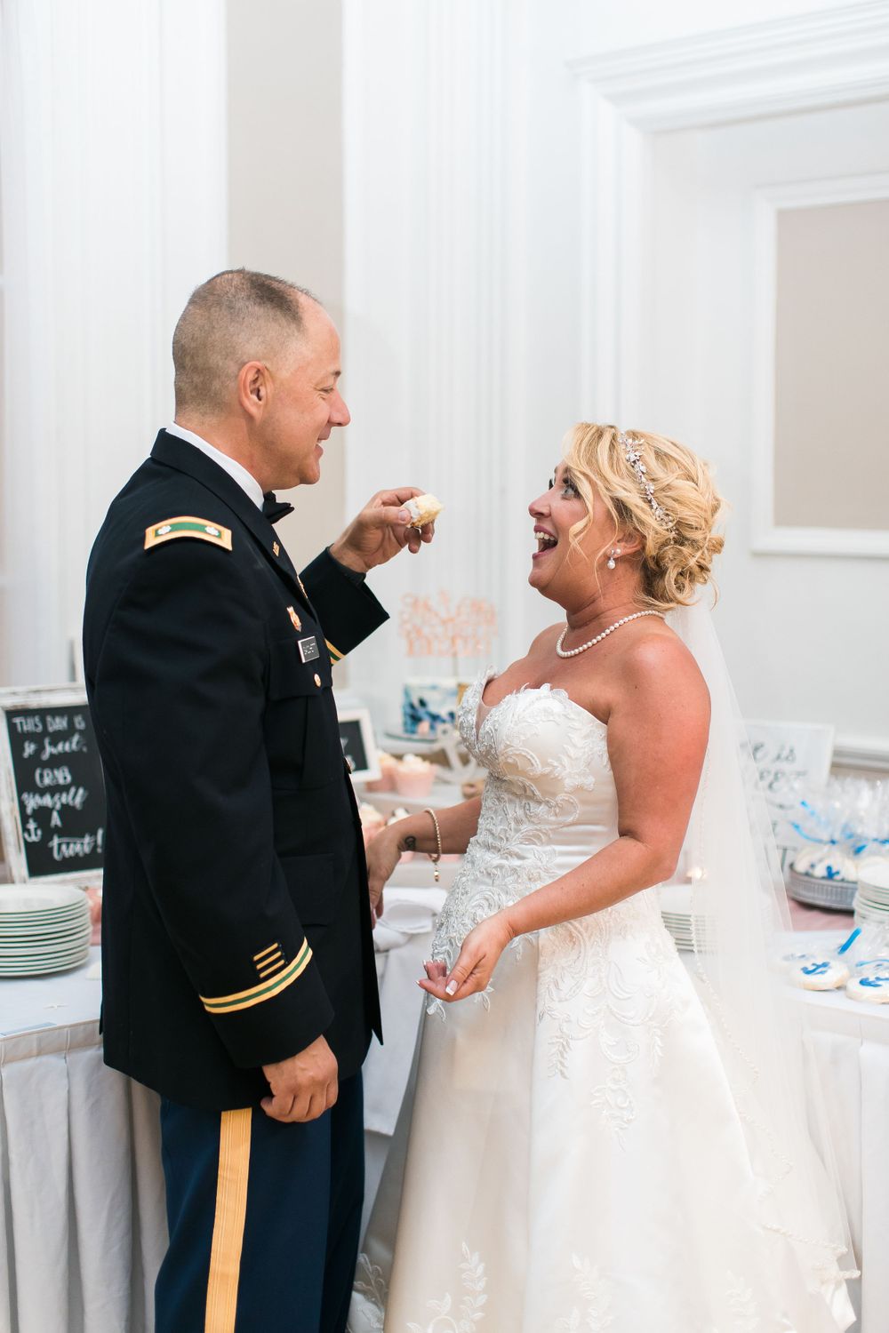 A wedding couple shares a joyful moment by a decorated reception table in a grand white room.