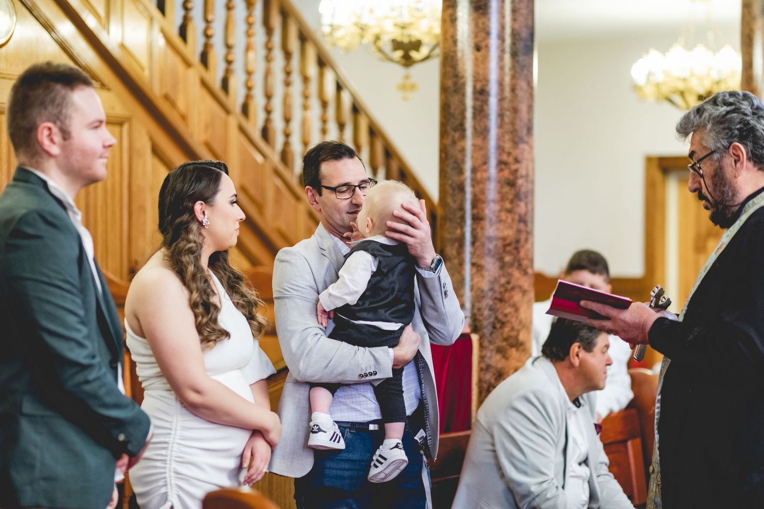 Religious ceremony participants gathered near marble columns and wooden staircase in church interior.