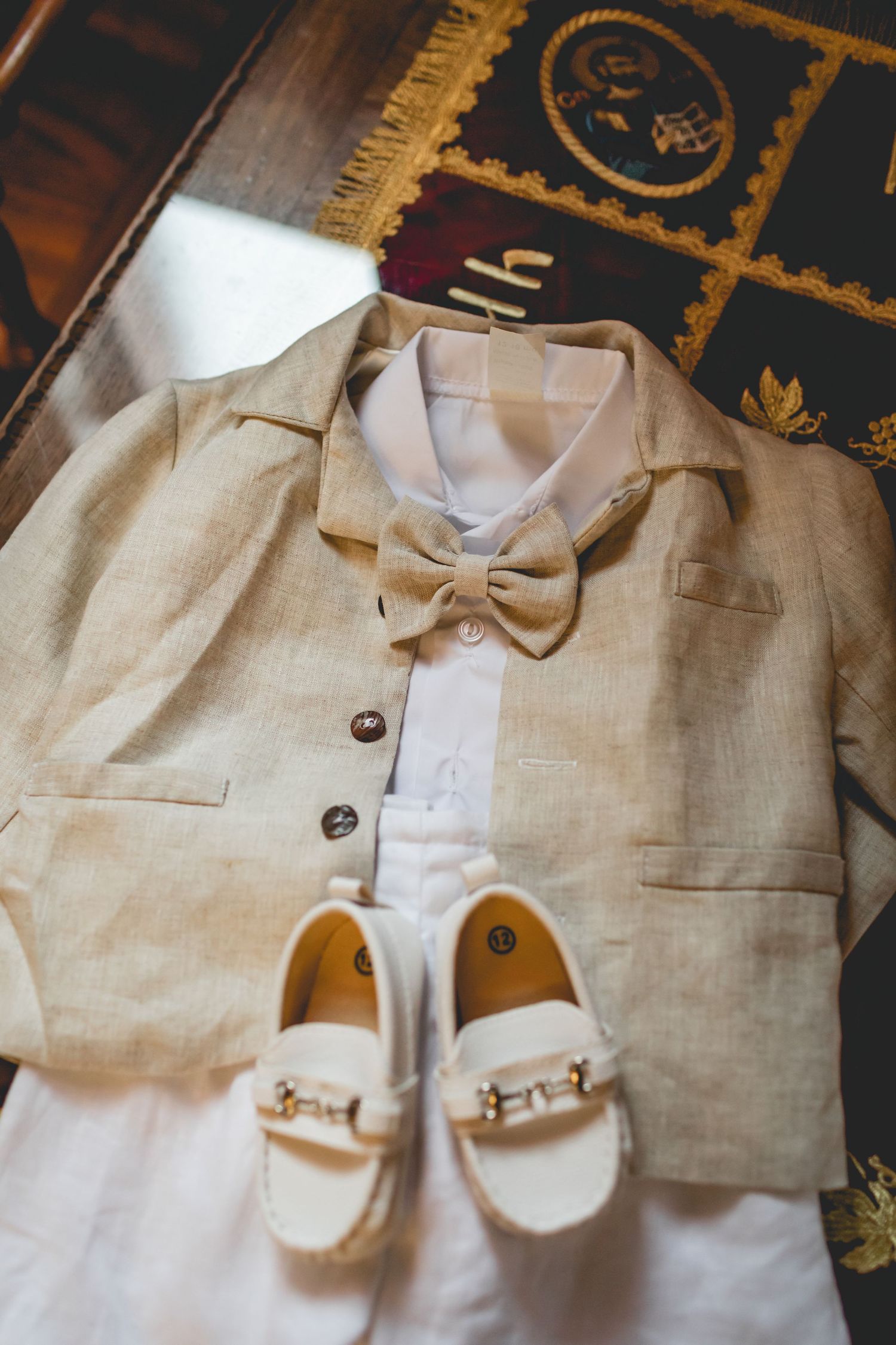 Formal child's outfit with beige jacket, white shirt, bow tie and tiny loafers laid flat.