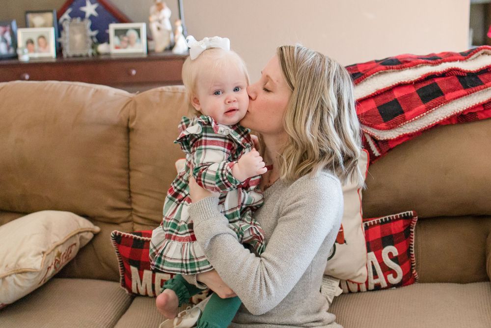 Mother in gray sweater kisses baby in plaid outfit on brown couch.
