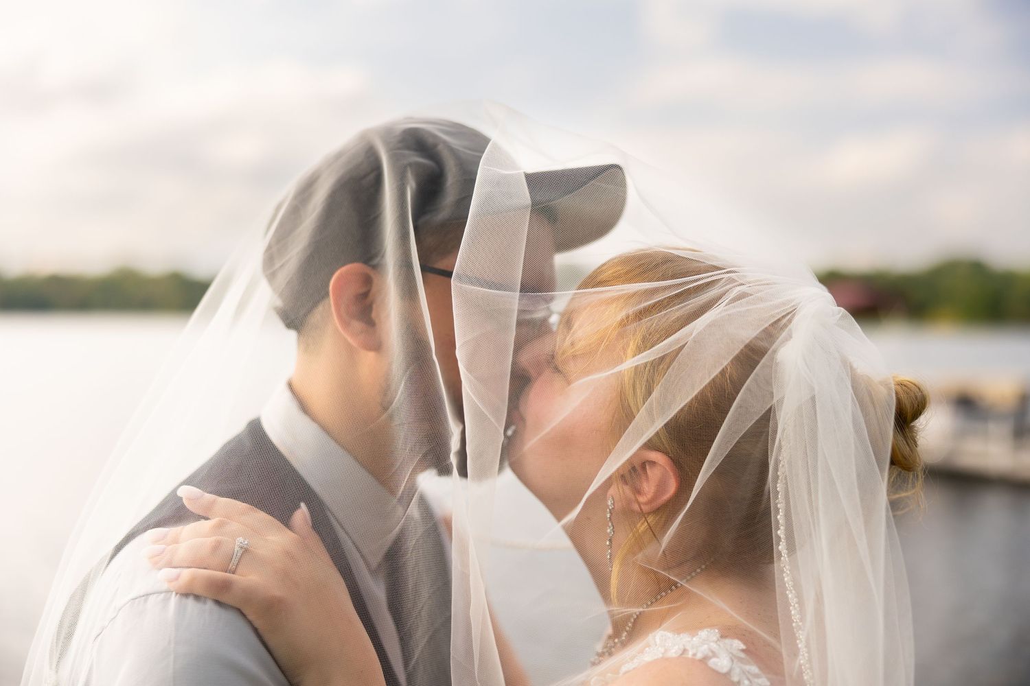Intimate wedding kiss captured through flowing white veil against waterfront backdrop.