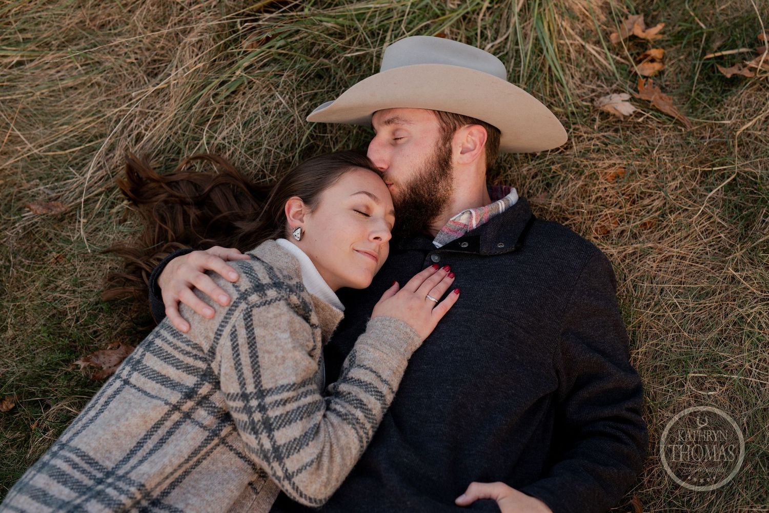 A couple shares an intimate moment while lying in tall grass with autumn trees in the background.