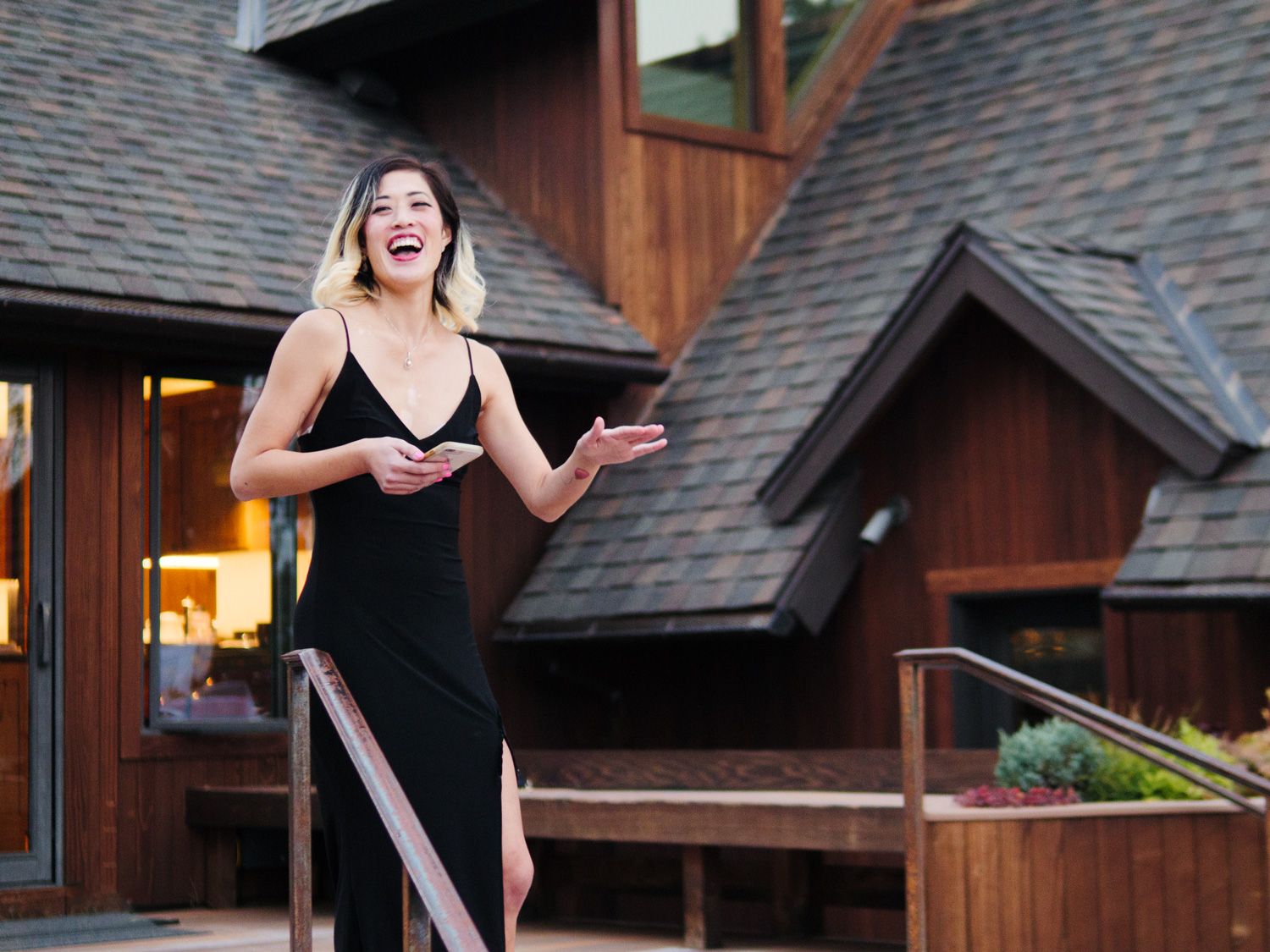 A woman in a black evening gown stands on wooden deck steps near a cabin-style building.