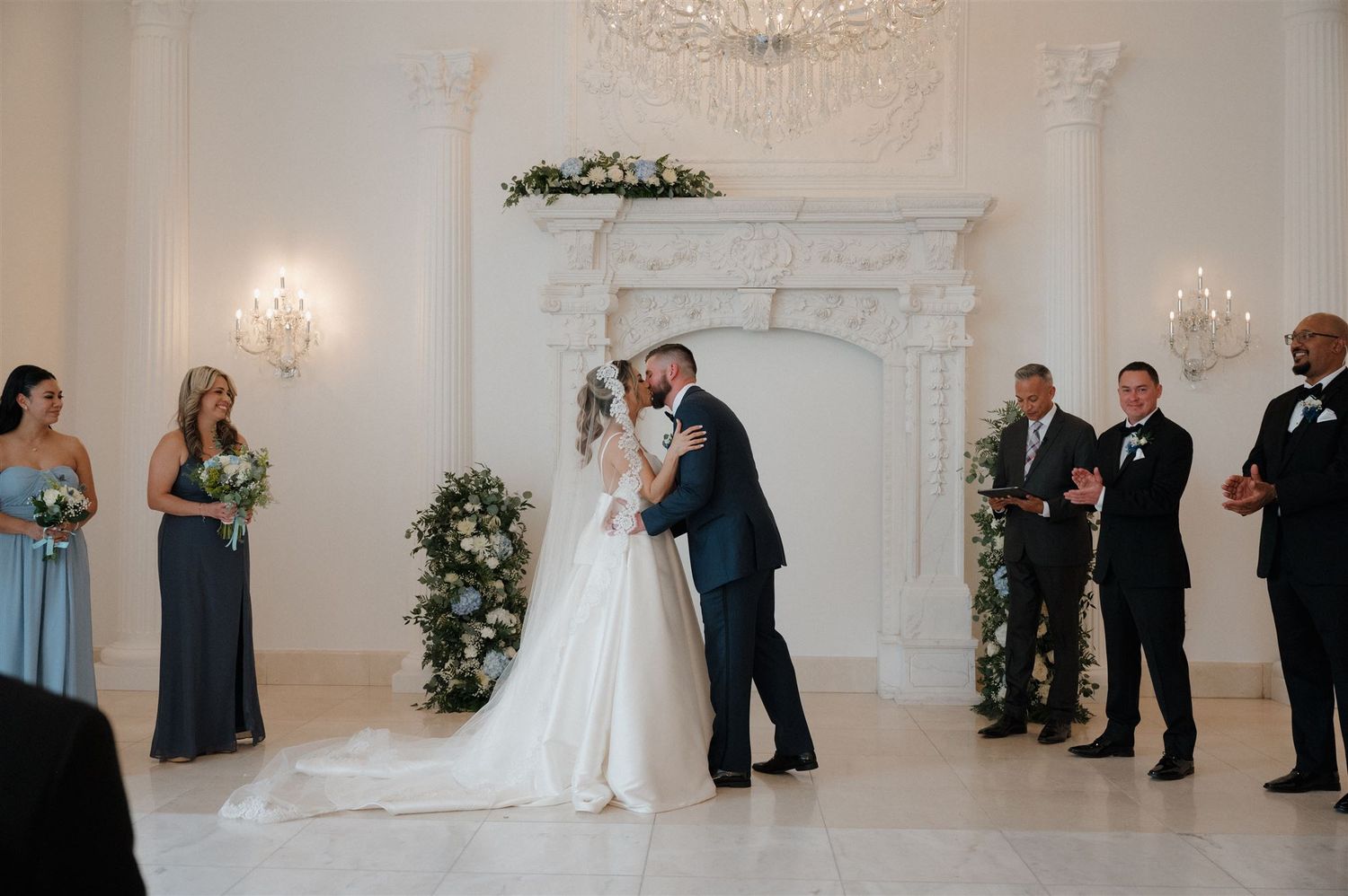 An intimate wedding ceremony unfolds beneath a grand chandelier in a classic white venue.