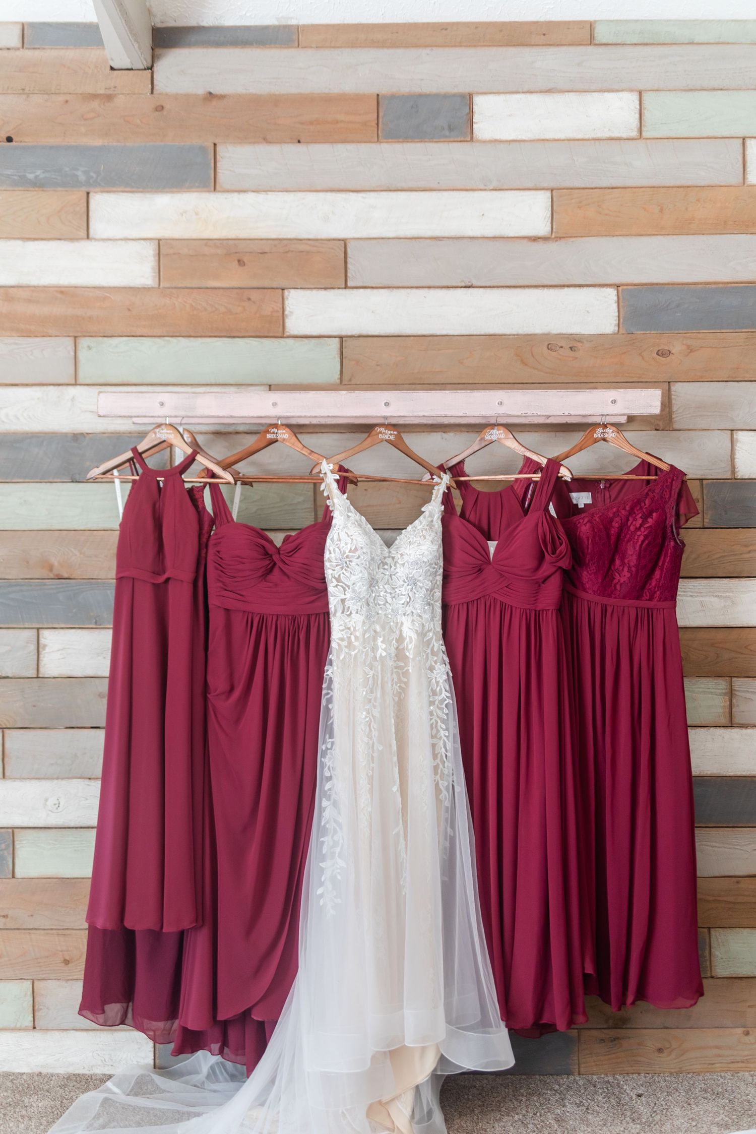 Burgundy bridesmaid dresses hanging beside a white wedding gown on a rustic wooden wall.