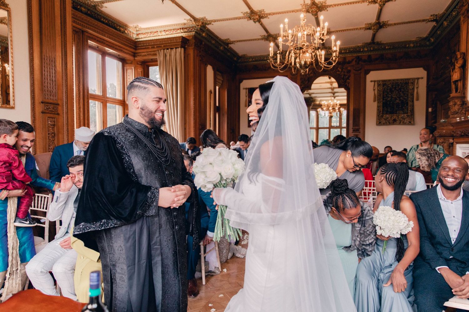 Wedding party celebrates in an ornate ballroom with elegant chandeliers and decorative woodwork.