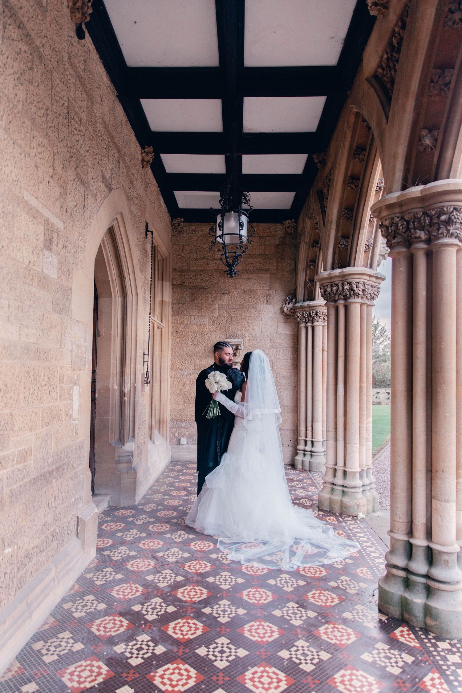 Wedding couple poses in gothic stone archway corridor with decorative tiled floor.