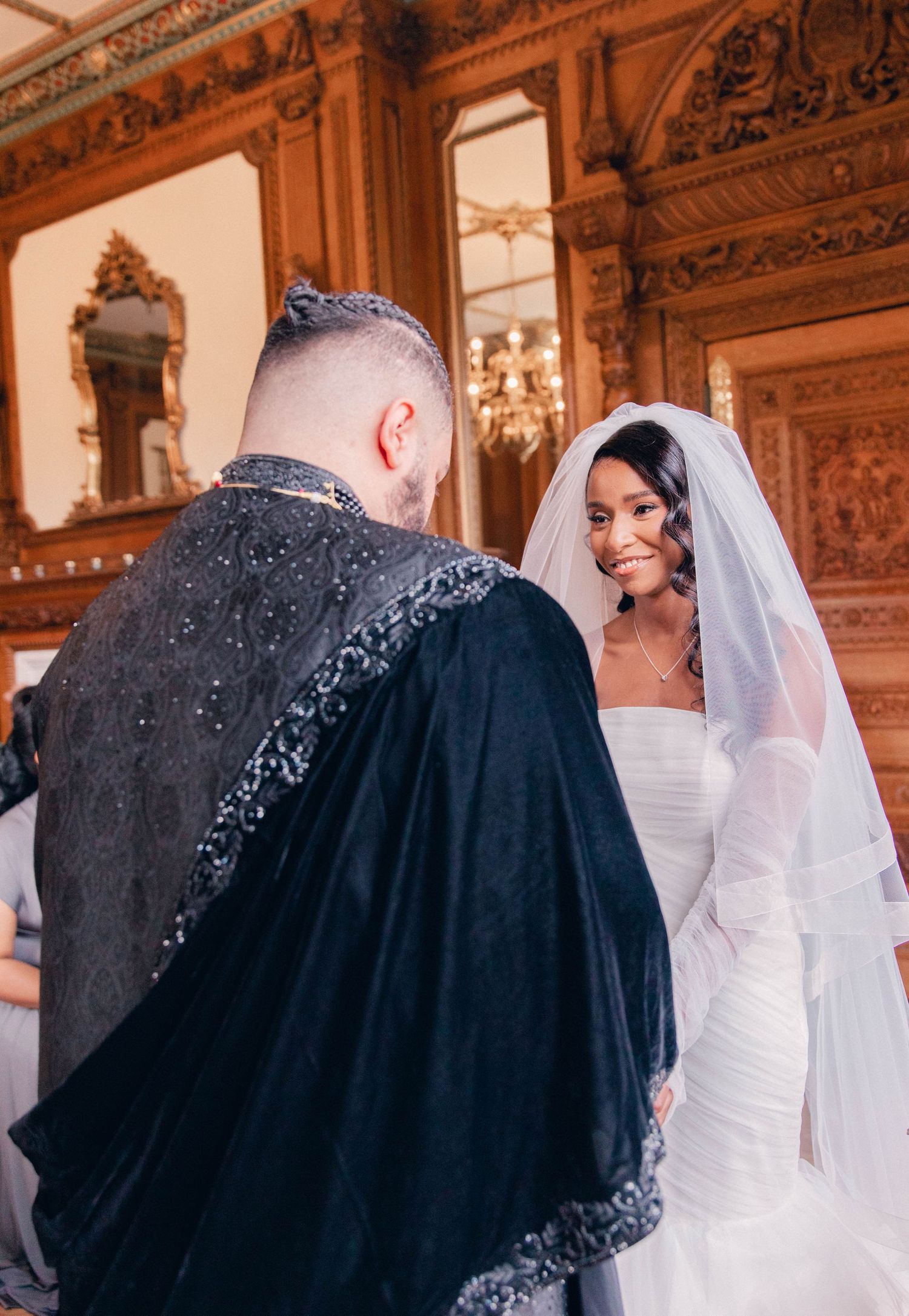 Bride in white dress stands with guests in an elegant wood-paneled room with ornate details.