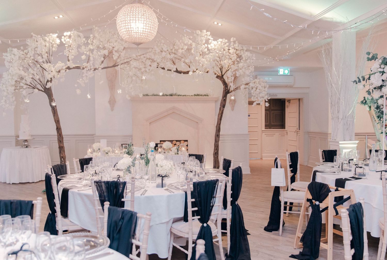 Elegant white wedding reception room decorated with crystal chandeliers and tree centerpieces.