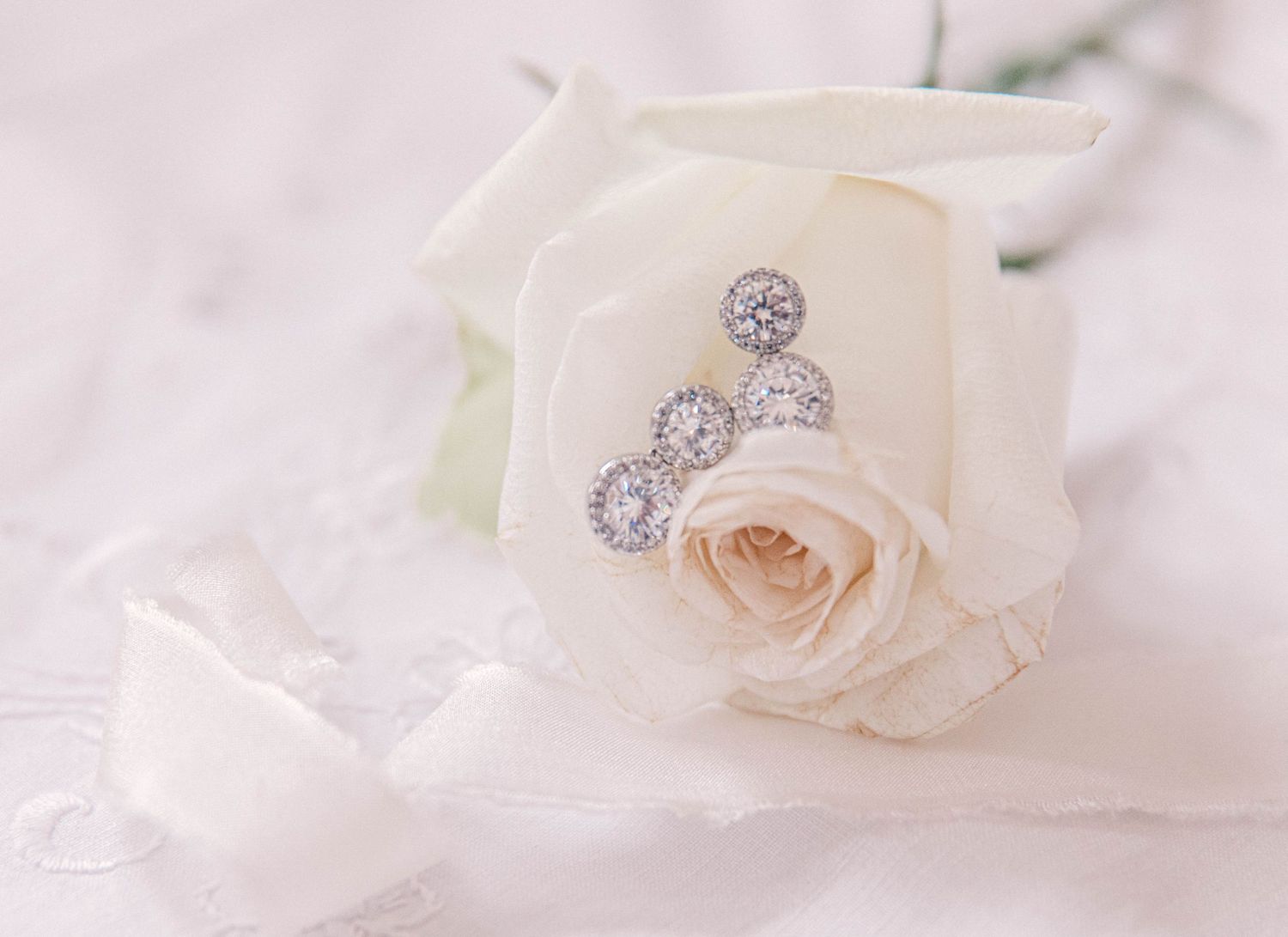 Delicate blush rose with sparkling diamond earrings on soft white fabric.