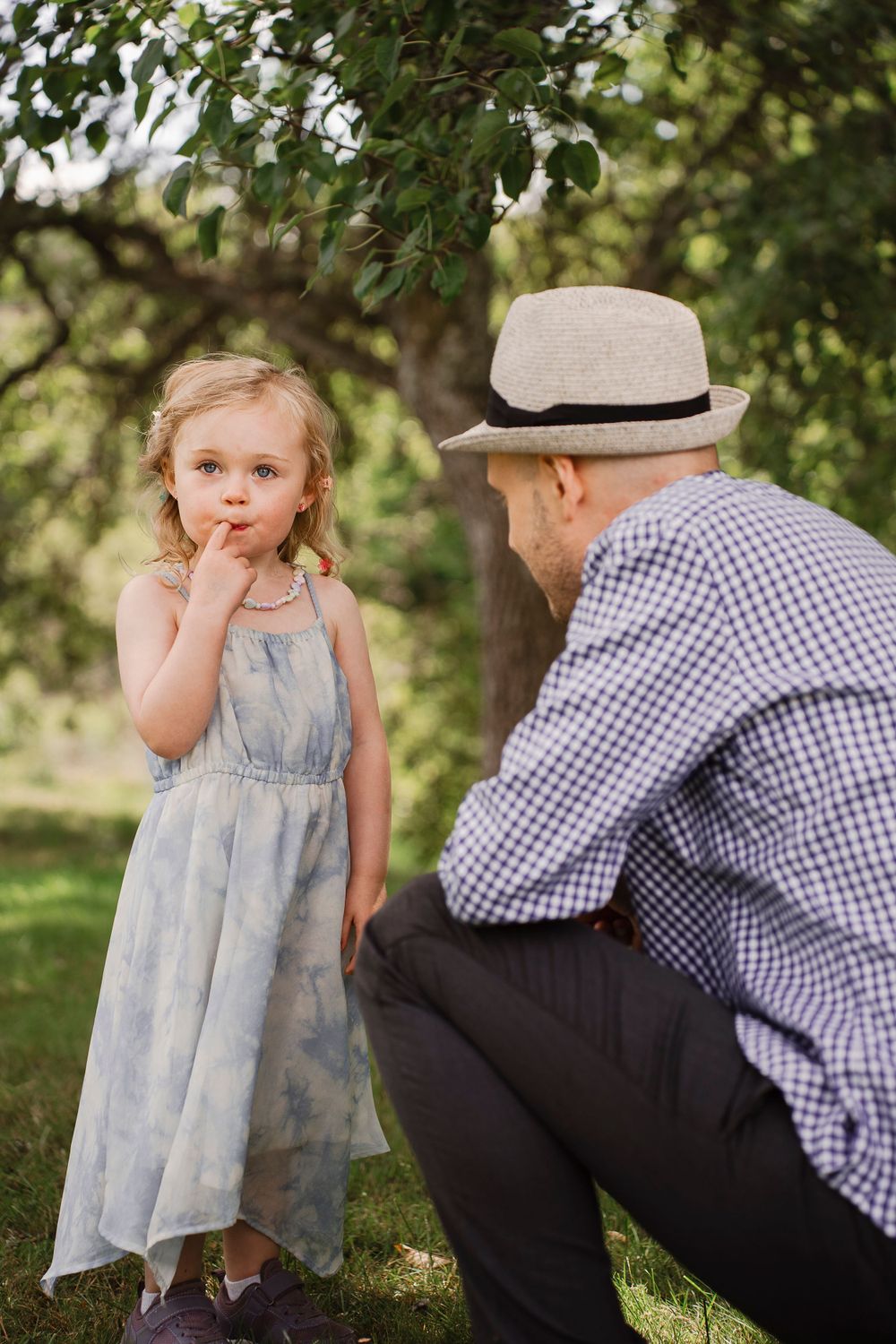 Tender moment between a young child in blue dress and adult in checkered shirt under trees.