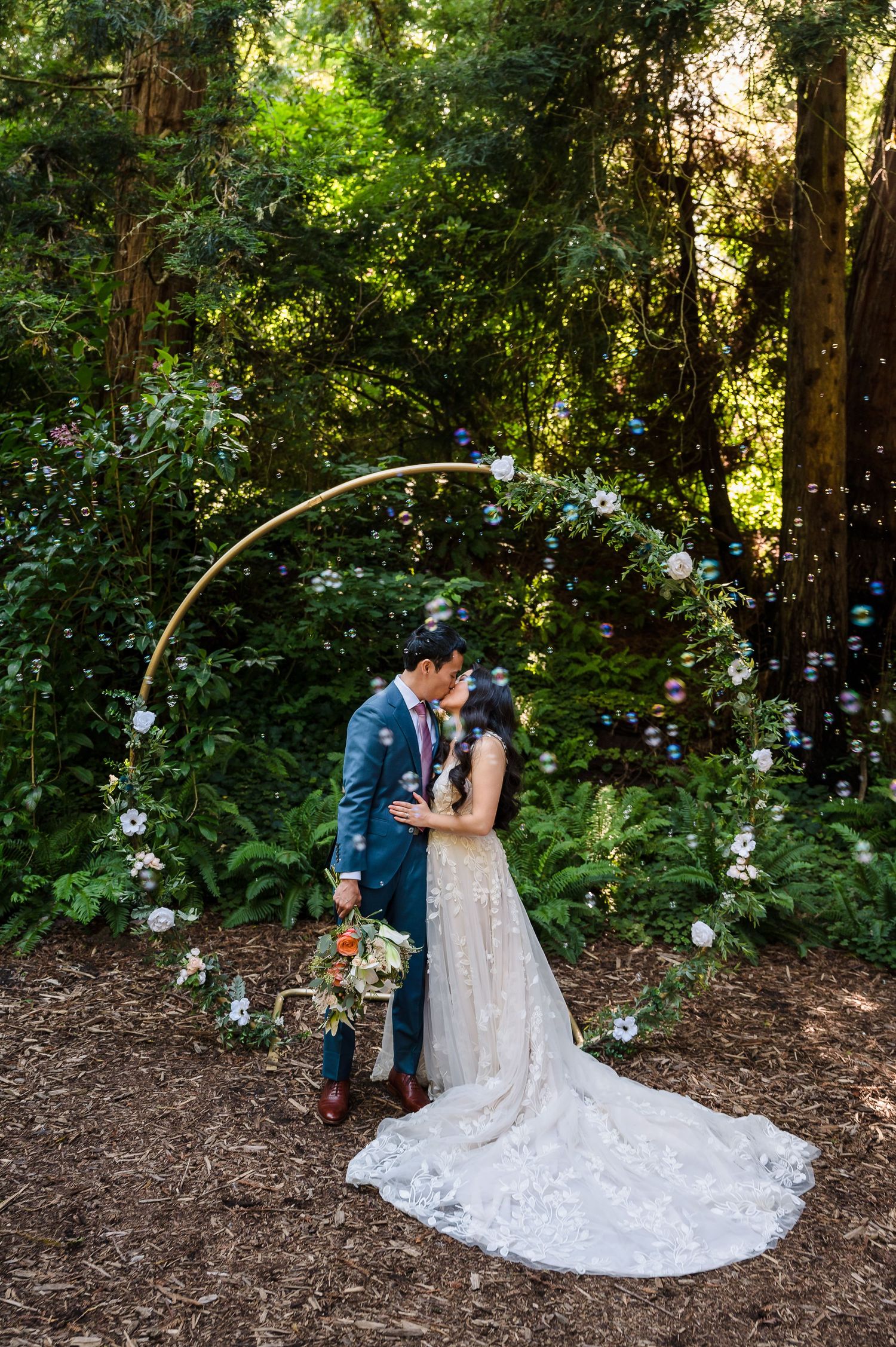 A couple kisses under a curved floral arch in a forest setting with floating bubbles.