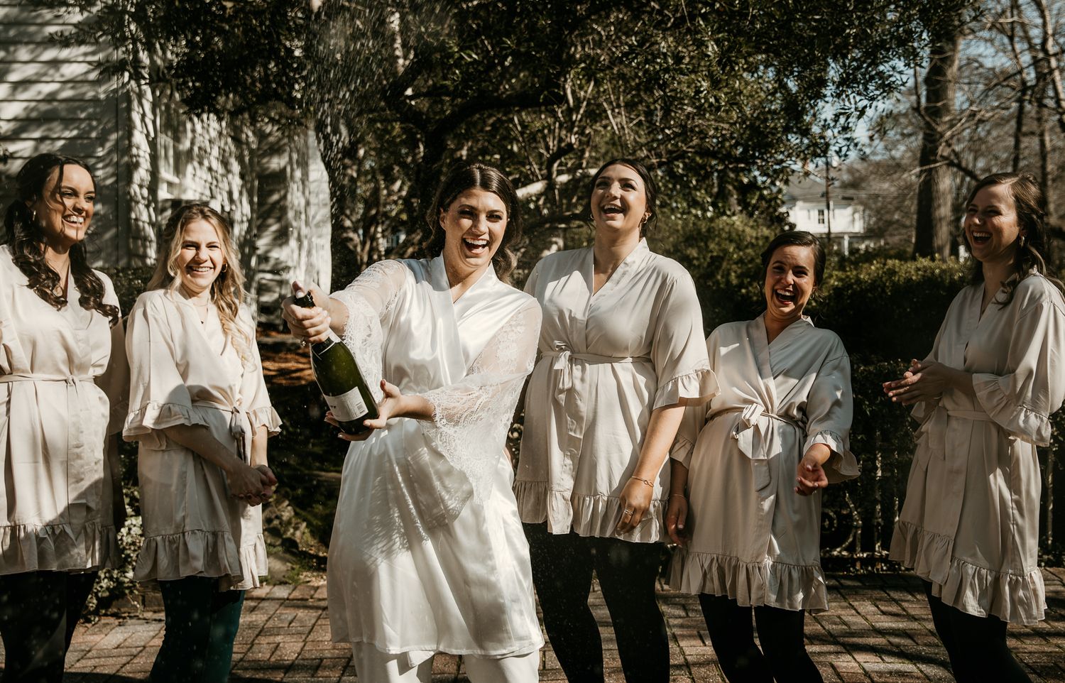 Wedding party in matching light-colored dresses celebrating on sunny patio.