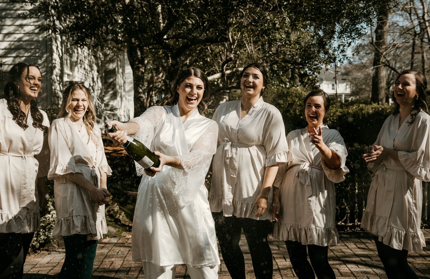 Bridal party in coordinated white and cream dresses laughing together outdoors.