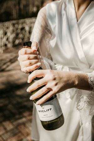 Close-up of hands in white robe opening wine bottle during wedding preparations.