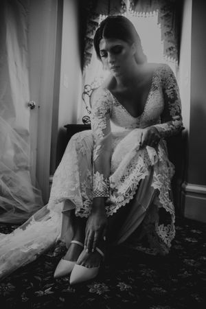 Bride in vintage-style lace wedding gown sitting on floral carpet, putting on white shoes in black and white photo.