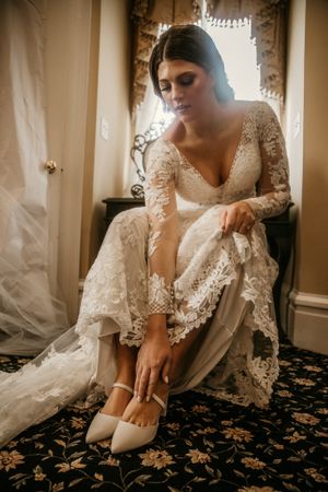 Bride in long sleeve lace wedding dress adjusting her shoes while sitting in a sunlit room.