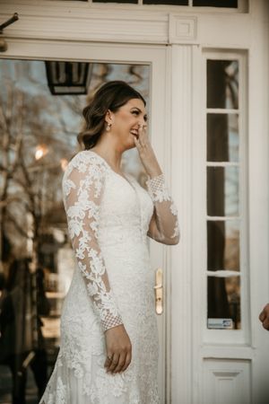 Elegant bride in long-sleeve lace wedding gown standing by white doorway.