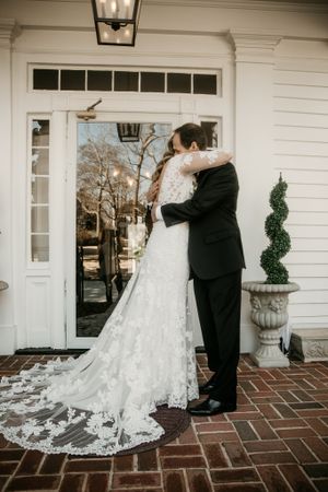 A wedding couple embraces on a brick porch in front of a white colonial-style entrance.