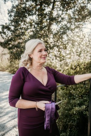 A woman in a purple dress stands outdoors against a natural backdrop.