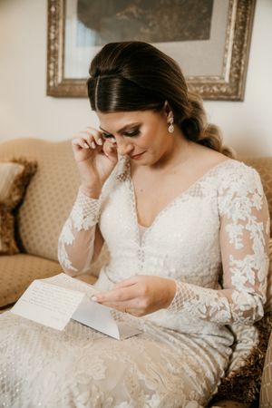 Emotional moment as bride in lace gown reads letter while seated on vintage sofa.