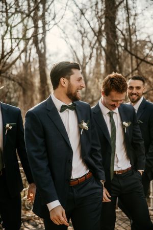 Groomsmen sharing a lighthearted moment on wedding day in navy suits against bare winter trees.