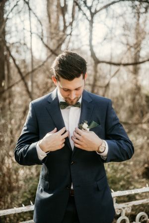 Groom in navy tuxedo adjusting boutonniere while standing outdoors in winter setting.