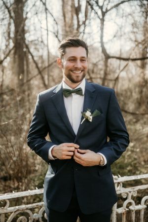 Groom in classic navy tuxedo smiling outdoors with winter foliage background.