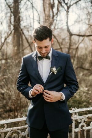 Groom adjusting bowtie on navy tuxedo against rustic winter backdrop.