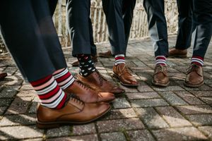 Groomsmen show off their matching brown leather dress shoes and red striped socks on stone pavement.