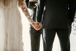 Couple holds hands during wedding ceremony with sunlight streaming through white lace dress.