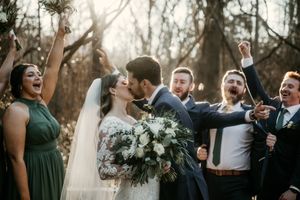 Wedding party celebrates joyfully with raised arms during an outdoor ceremony in a wooded setting.