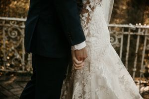 Close-up detail of hands and lace wedding dress against groom's black suit during ceremony.