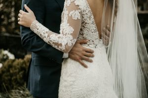 Detail shot of lace wedding dress and groom's suit as couple embraces during ceremony.