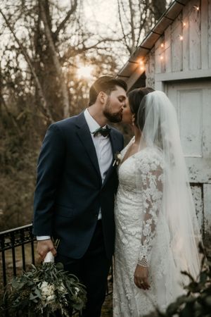 Romantic sunset wedding portrait against rustic barn with string lights and greenery.