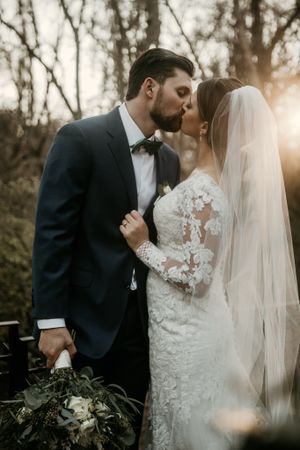Intimate sunset wedding portrait with couple holding greenery bouquet against wooded backdrop.