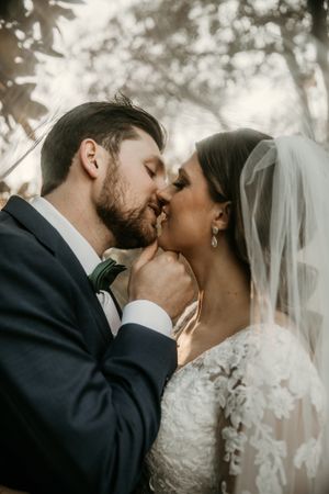 Intimate wedding portrait showing a tender kiss between newlyweds outdoors with soft lighting and veil details.