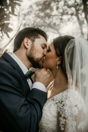 Romantic wedding portrait showing an intimate kiss between newlyweds with sunlit forest backdrop.