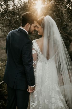 Backlit wedding portrait showing intimate moment between newlyweds with sun flare through trees.