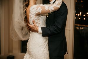 Close-up wedding detail showing embrace with lace dress and classic tuxedo indoors with twinkle lights.
