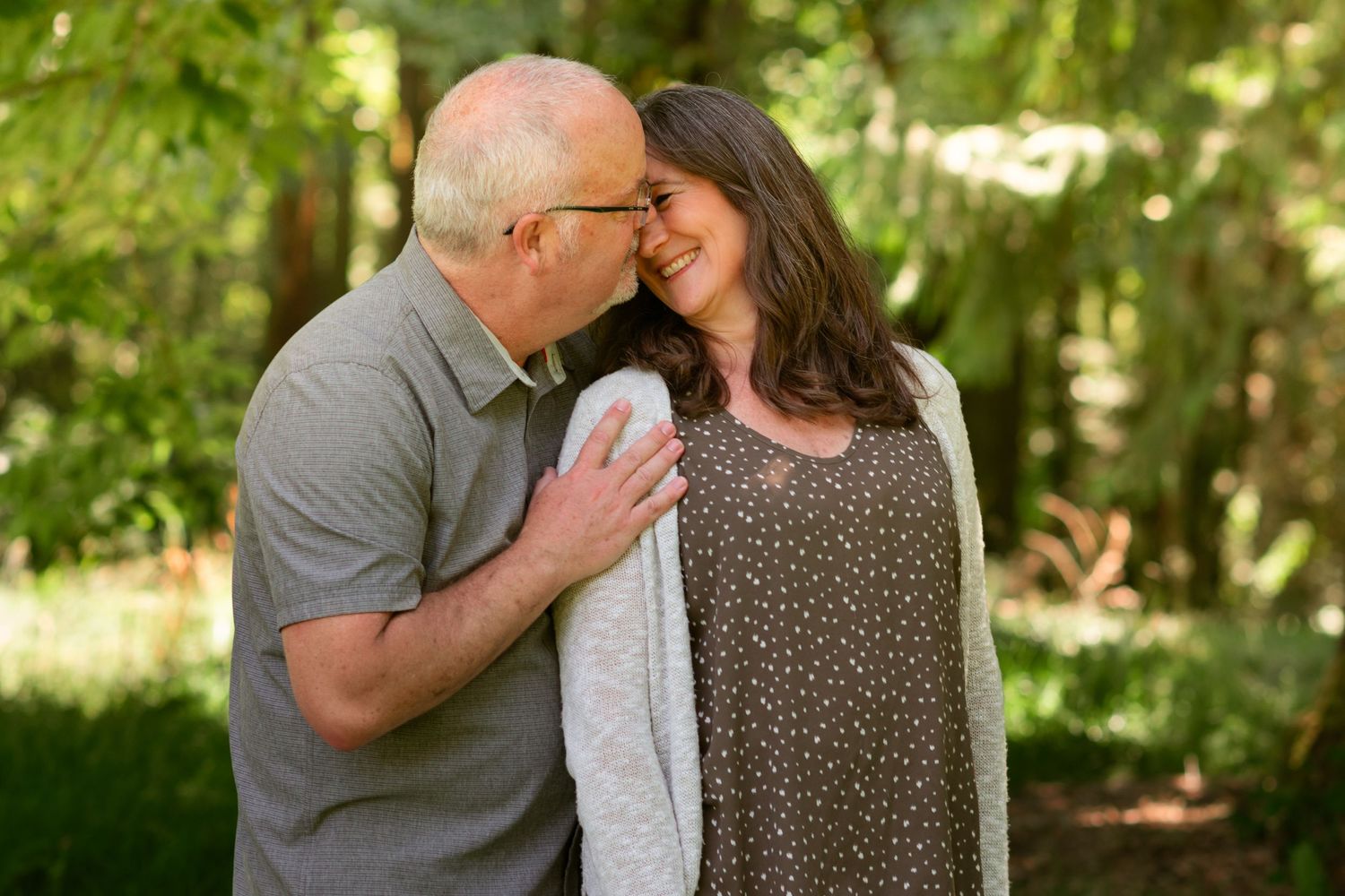 husband and wife smiling close to each other's faces. photo taken in a treed area.