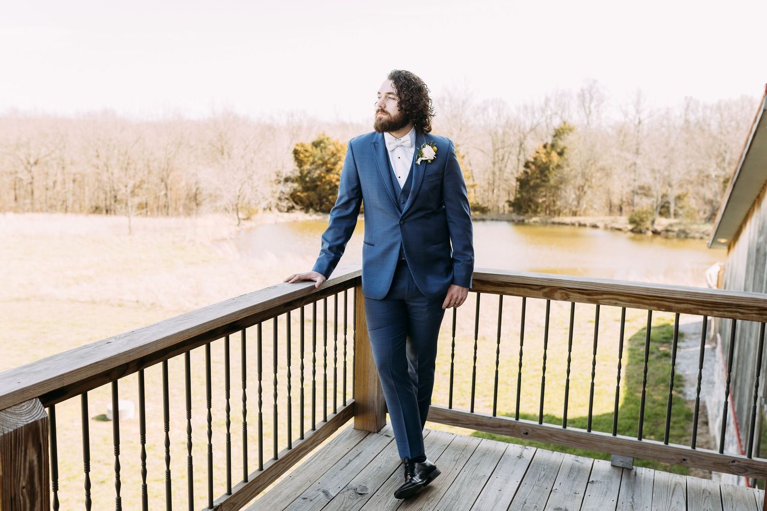 Groom staring off a second floor outdoor balcony looking out into a beautiful treelined field
