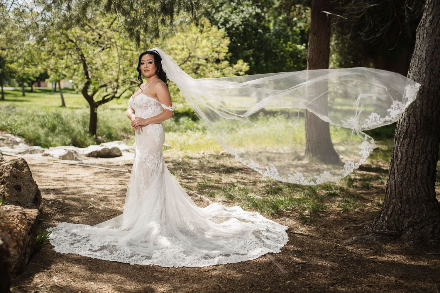 Bride with flowing veil poses in natural woodland setting wearing fitted lace gown.