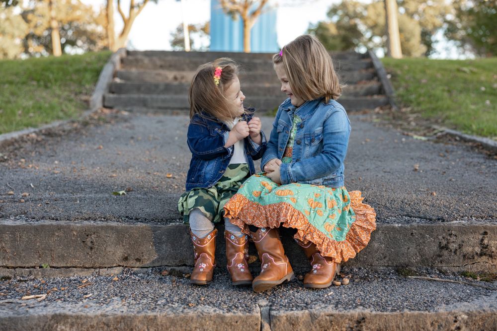 Two small children sit together on concrete steps wearing matching boots and outfits.
