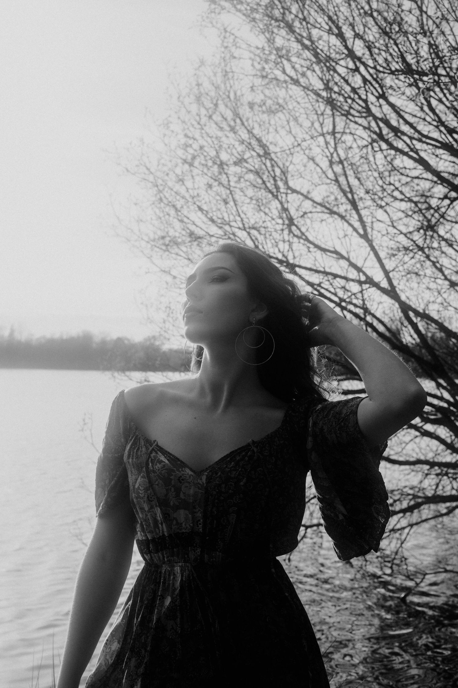 A dramatic black and white portrait of a woman in a dress posing by a lake with bare trees.