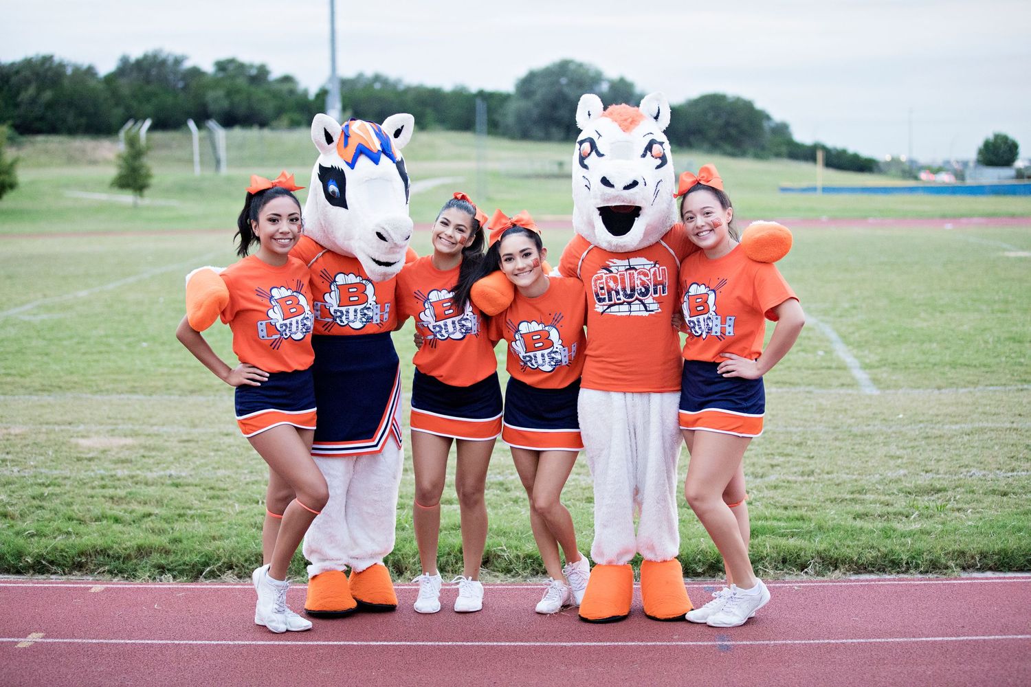 Cheerleaders in orange uniforms pose with mascots wearing white tiger costumes on an athletic track.