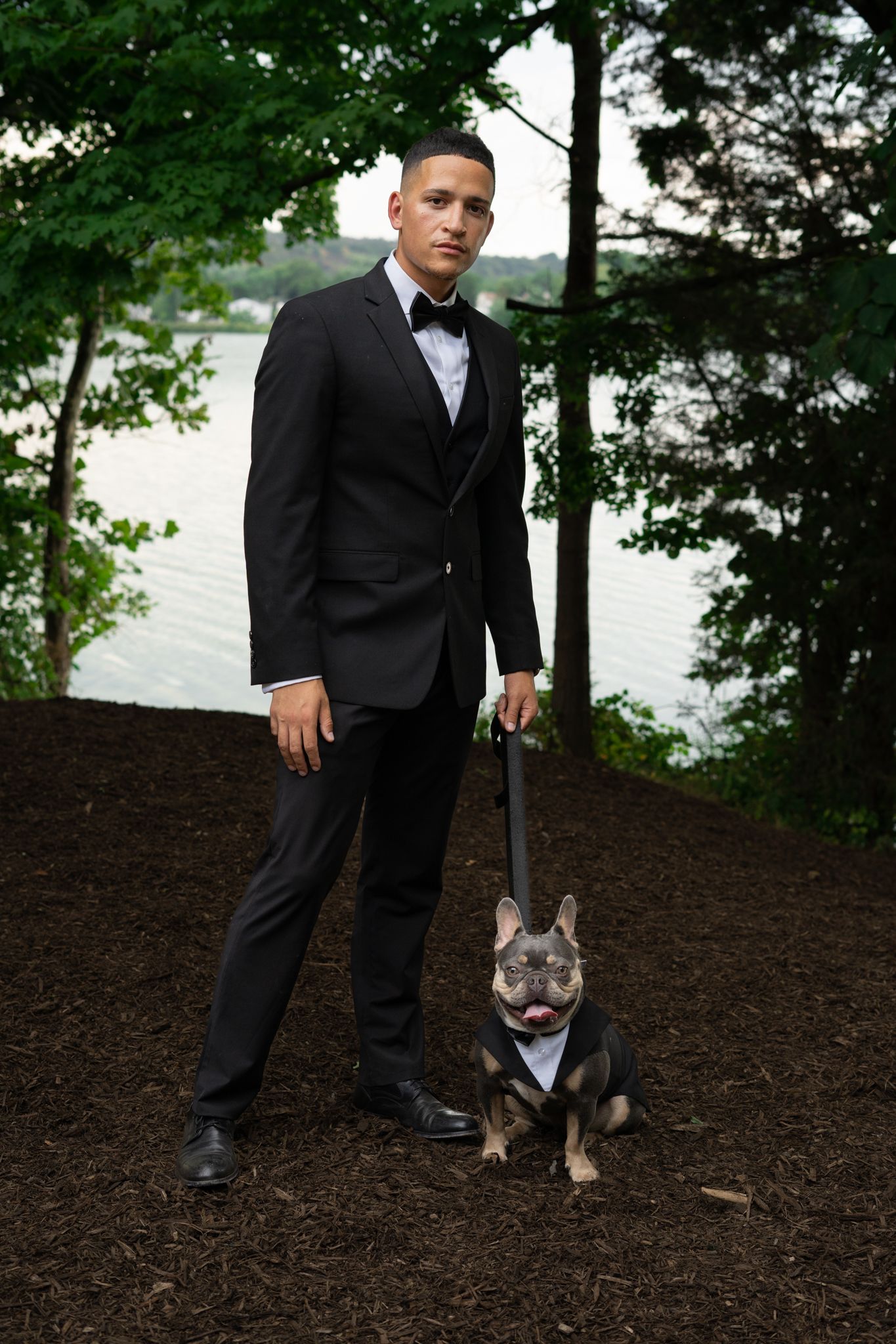 Dog accompanies formally dressed people walking through a wooded area near water.