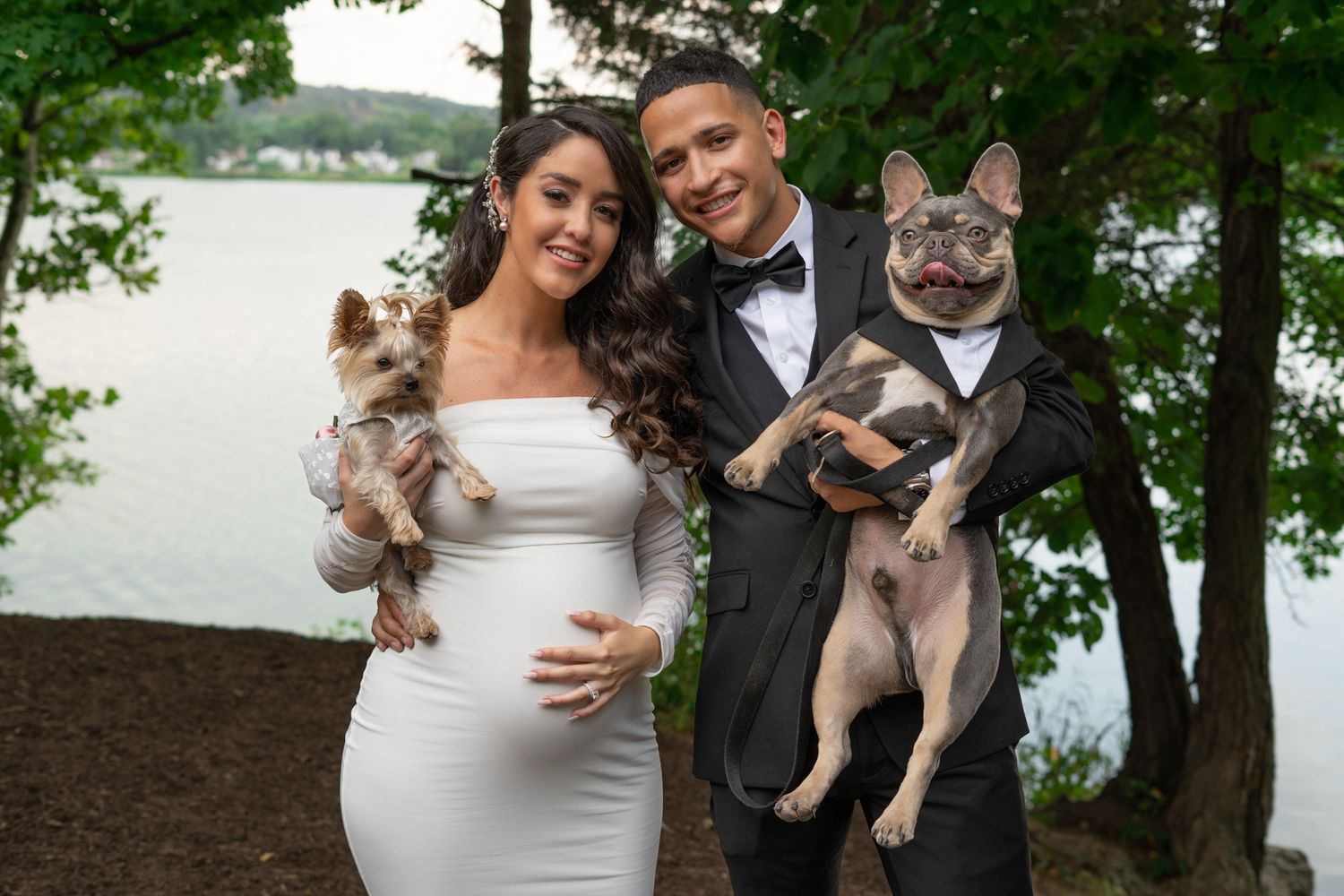 Couple in formal attire holding small dogs outdoors by a lake.
